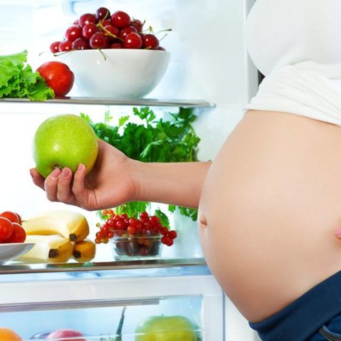 Kobieta w ciąży stoi przy lodówce i trzyma w ręku jabłko