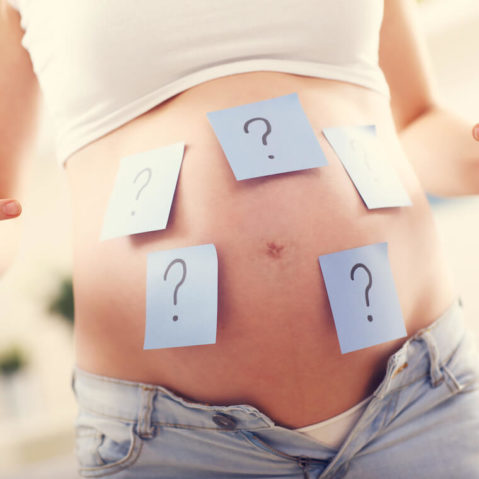 Kadr na brzuch kobiety w ciąży. Na brzuchu znajdują się przyklejone kartki ze znakami zapytania.