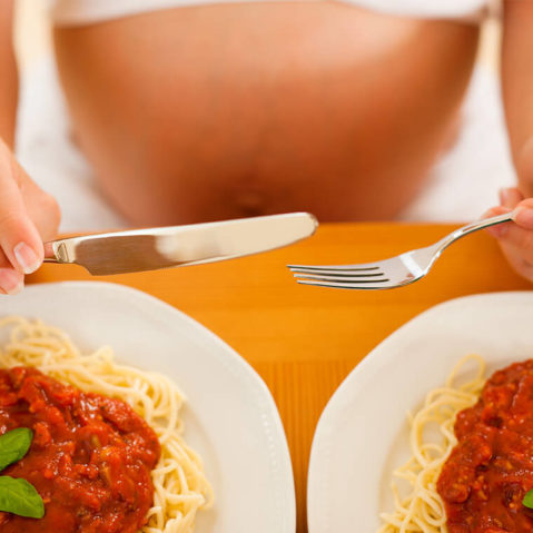 Kobieta w ciąży siedzi z widelcem i nożem w ręce. Przed nią znajdują się dwa talerze ze spaghetti.