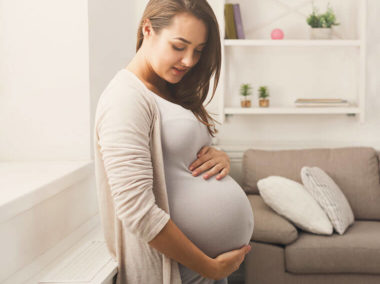 Kobieta w ciąży opiera się o parapet. Dłonie ma ułożone na brzuchu.