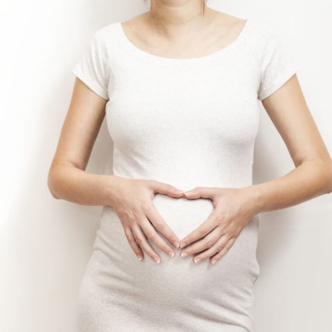 Kobieta w ciąży opiera się o ścianę. Na brzuchu trzyma ręce ułożone w kształt serca.